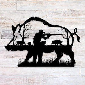 Puede incluir: Arte de pared de metal negro que representa a un cazador apuntando con un rifle a un jabalí. El diseño de silueta incluye el contorno del jabalí, árboles, hierba y jabalíes más pequeños, creando una escena de caza. La obra de arte está montada sobre un fondo de madera blanca.