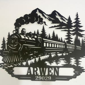 May include: A black metal wall art piece featuring a steam locomotive train travelling through a forest with mountains in the background. The design includes a rectangular nameplate at the bottom with the text ARWEN and the number 29029.