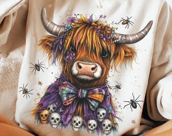 May include: A cream-coloured sweatshirt with a Highland cow illustration. The cow has a floral crown, a purple scarf with a bow, and skulls. Black spiders are scattered around the cow. The design is whimsical and colourful.
