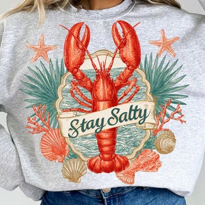 May include: A light gray sweatshirt featuring a large red lobster graphic with the words "Stay Salty" on a banner. The design includes starfish, seashells, and coral, evoking a coastal theme.