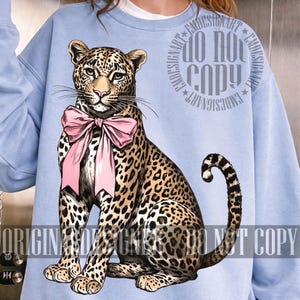 May include: Light blue sweatshirt featuring a detailed illustration of a leopard wearing a pink bow. The leopard has a spotted coat and is sitting upright. The sweatshirt has the text "DO NOT COPY" and "ORIGINAL DESIGNS" printed on it.