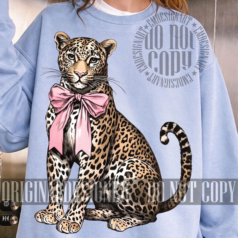 May include: Light blue sweatshirt featuring a detailed illustration of a leopard wearing a pink bow. The leopard has a spotted coat and is sitting upright. The sweatshirt has the text "DO NOT COPY" and "ORIGINAL DESIGNS" printed on it.