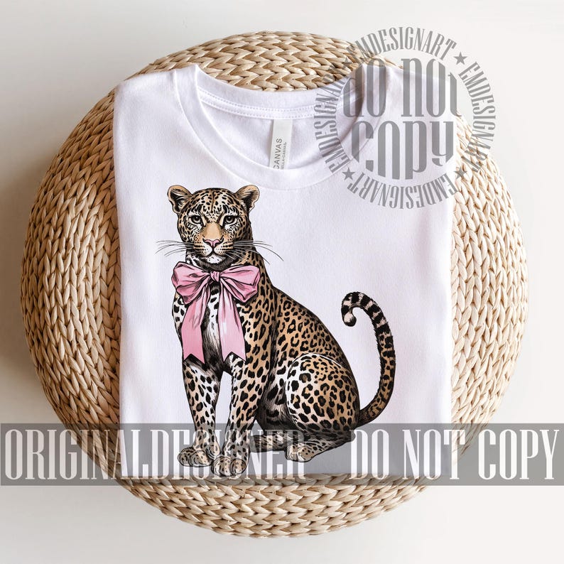 May include: White t-shirt featuring a seated leopard illustration with a pink bow. The leopard has a spotted coat and a curled tail. The shirt is on a woven, circular surface. The text "ORIGINAL DESIGNART DO NOT COPY" is printed on the bottom.