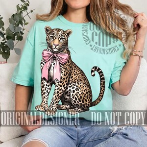 May include: A light blue t-shirt featuring a detailed illustration of a leopard wearing a pink bow. The leopard has a spotted coat and is sitting upright. The text "ORIGINAL DESIGNER DO NOT COPY" is printed at the bottom.