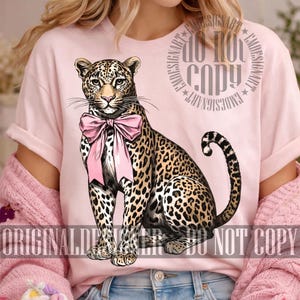 May include: A light pink t-shirt featuring a detailed illustration of a leopard wearing a pink bow. The leopard has a spotted coat and is sitting upright. The words "ORIGINAL DESIGN" and "DO NOT COPY" are printed on the shirt.