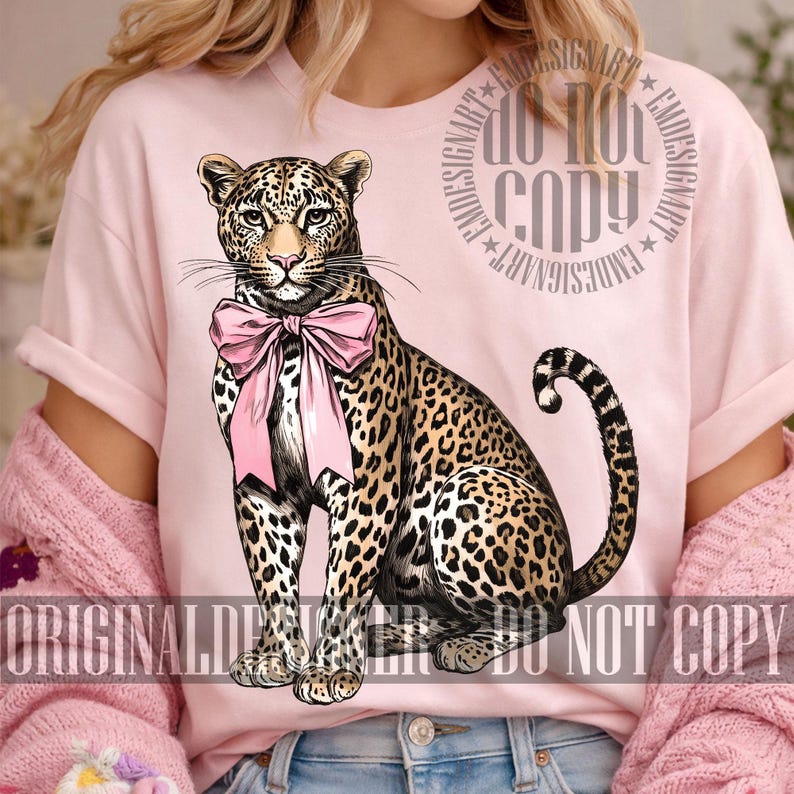 May include: A light pink t-shirt featuring a detailed illustration of a leopard wearing a pink bow. The leopard has a spotted coat and is sitting upright. The words "ORIGINAL DESIGN" and "DO NOT COPY" are printed on the shirt.