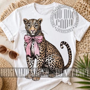 May include: White t-shirt featuring a detailed illustration of a leopard with a pink bow. The leopard has a spotted coat and is sitting upright. The text "ORIGINAL DESIGN" and "DO NOT COPY" are visible.