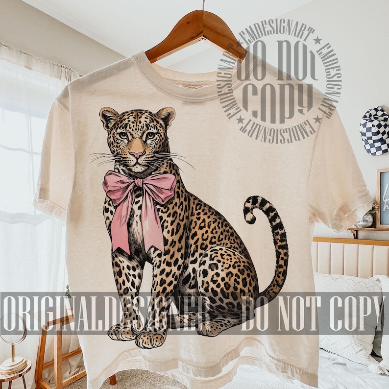 May include: Cream-colored t-shirt featuring a detailed illustration of a leopard with a pink bow. The leopard has a spotted coat and is sitting upright. The shirt is hanging on a wooden hanger.