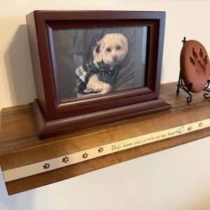 May include: A wooden shelf with a framed photo of a dog, a paw print keepsake, and the inscription "Dogs leave paw prints on our hearts." The shelf is made of dark wood with a light wood accent.