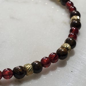 May include: A close-up of a red and brown beaded bracelet with gold accents. The bracelet is made of small, round beads that are strung together on a thin cord.