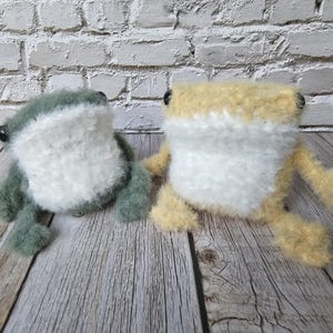 May include: Two handmade plush frog toys. One is green and white, the other is yellow and white. Both have black button eyes and are sitting on a wooden surface. The frogs are soft and fluffy, with a whimsical design.