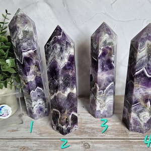 K&ouml;nnte beinhalten: Vier Amethyst-Kristall-Obelisken mit lila, wei&szlig;en und grauen Mustern. Die Kristalle stehen auf einer Holzoberfl&auml;che. Die Obelisken haben eine polierte, glatte Oberfl&auml;che und eine spitze Spitze.