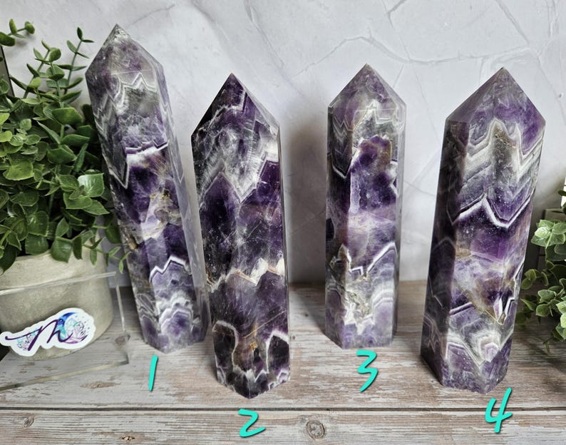 K&ouml;nnte beinhalten: Vier Amethyst-Kristall-Obelisken mit lila, wei&szlig;en und grauen Mustern. Die Kristalle stehen auf einer Holzoberfl&auml;che. Die Obelisken haben eine polierte, glatte Oberfl&auml;che und eine spitze Spitze.
