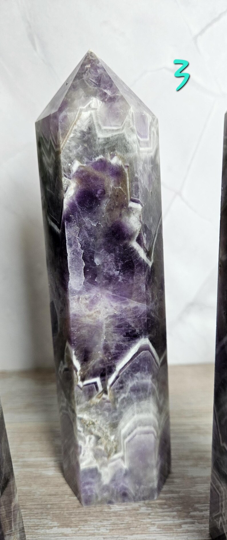 K&ouml;nnte beinhalten: Ein hoher, facettierter Amethyst-Kristallpunkt mit polierter Oberfl&auml;che. Der Kristall weist eine Mischung aus violetten, wei&szlig;en und grauen Farbt&ouml;nen mit sichtbaren inneren Mustern auf. Der Kristall ist ein dekorativer Gegenstand.