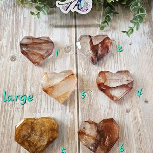 Puede incluir: Seis cristales de cuarzo claro en forma de coraz&oacute;n con inclusiones marrones y doradas. Los cristales est&aacute;n dispuestos en dos filas de tres. Los cristales est&aacute;n etiquetados con los n&uacute;meros del 1 al 6. La palabra "grande" est&aacute; escrita debajo de los cristales.