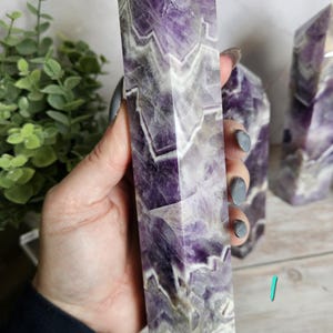 K&ouml;nnte beinhalten: Ein hoher, rechteckiger Amethyst-Kristallpunkt mit facettierter Spitze. Der Kristall weist eine Mischung aus violetten und wei&szlig;en B&auml;ndern auf. Ein weiterer Kristall ist im Hintergrund sichtbar. Der Kristall wird in einer Hand gehalten.