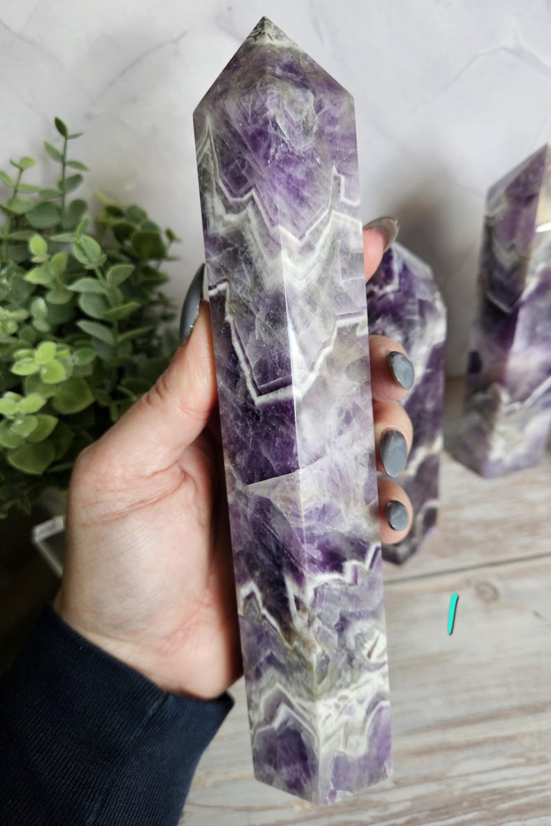 K&ouml;nnte beinhalten: Ein hoher, rechteckiger Amethyst-Kristallpunkt mit facettierter Spitze. Der Kristall weist eine Mischung aus violetten und wei&szlig;en B&auml;ndern auf. Ein weiterer Kristall ist im Hintergrund sichtbar. Der Kristall wird in einer Hand gehalten.