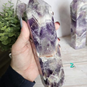 K&ouml;nnte beinhalten: Ein gro&szlig;er Amethyst-Kristallpunkt mit polierter, facettierter Oberfl&auml;che. Der Kristall weist eine Mischung aus tiefvioletten und wei&szlig;en Farbt&ouml;nen mit nat&uuml;rlichen Farb- und Texturvariationen auf. Der Kristall wird in einer Hand gehalten.