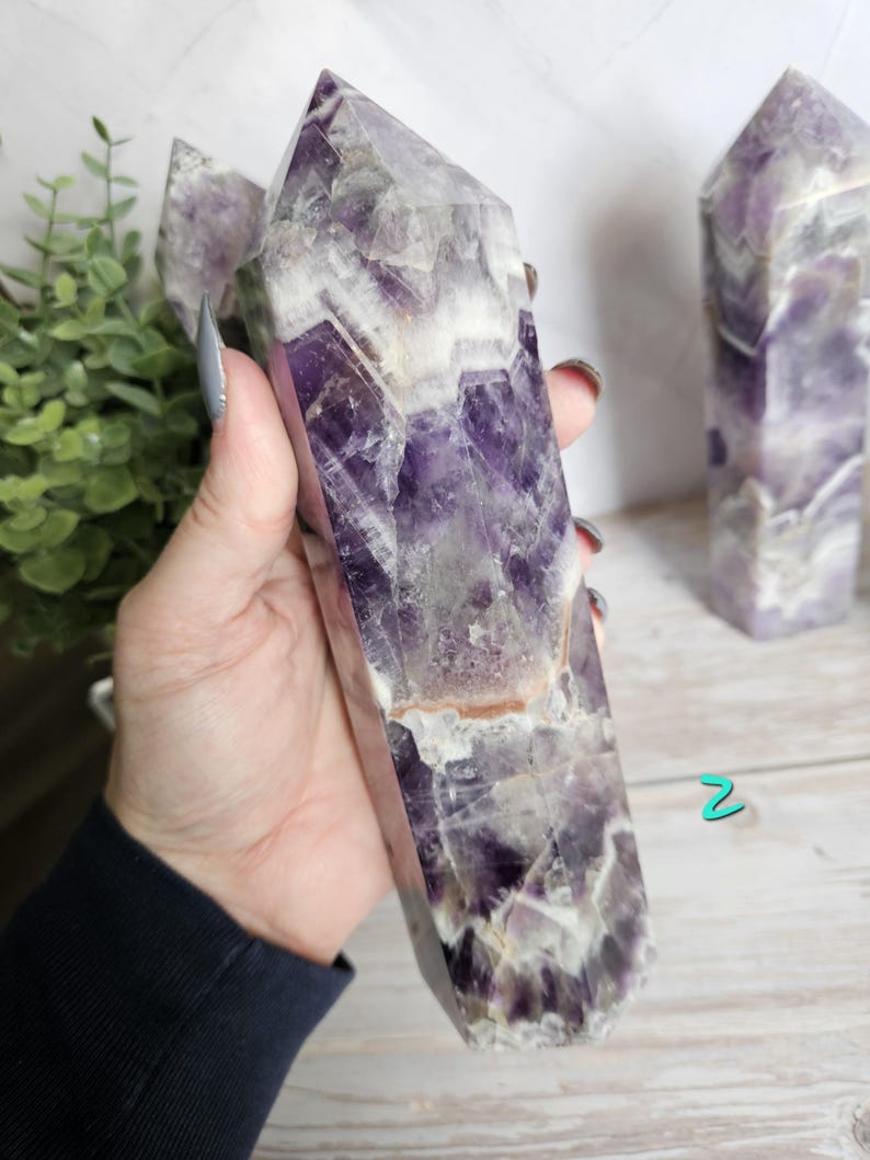 K&ouml;nnte beinhalten: Ein gro&szlig;er Amethyst-Kristallpunkt mit polierter, facettierter Oberfl&auml;che. Der Kristall weist eine Mischung aus tiefvioletten und wei&szlig;en Farbt&ouml;nen mit nat&uuml;rlichen Farb- und Texturvariationen auf. Der Kristall wird in einer Hand gehalten.