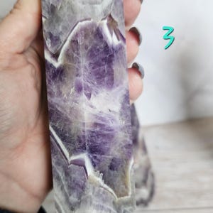 K&ouml;nnte beinhalten: Ein hoher, rechteckiger Amethyst-Kristallpunkt mit polierter Oberfl&auml;che. Der Kristall weist eine Mischung aus tiefvioletten und wei&szlig;en B&auml;ndern auf, die ein auff&auml;lliges visuelles Muster erzeugen. Die Spitze hat eine pyramidenf&ouml;rmige Form. Ein zweiter Kristall ist im Hintergrund zu sehen.