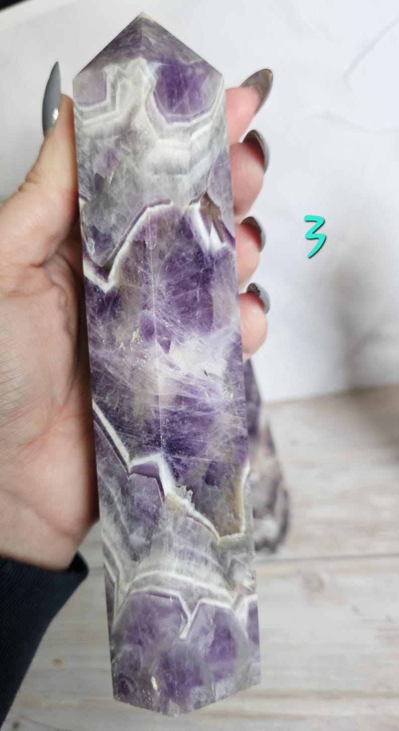 K&ouml;nnte beinhalten: Ein hoher, rechteckiger Amethyst-Kristallpunkt mit polierter Oberfl&auml;che. Der Kristall weist eine Mischung aus tiefvioletten und wei&szlig;en B&auml;ndern auf, die ein auff&auml;lliges visuelles Muster erzeugen. Die Spitze hat eine pyramidenf&ouml;rmige Form. Ein zweiter Kristall ist im Hintergrund zu sehen.
