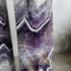 K&ouml;nnte beinhalten: Zwei Amethyst-Obelisken mit poliertem Finish. Der violett-wei&szlig; geb&auml;nderte Stein hat eine kristalline Struktur und ein geometrisches Muster. Die Obelisken sind rechteckig mit einer spitzen Spitze. Die Zahl "4" ist in T&uuml;rkis im Hintergrund geschrieben.
