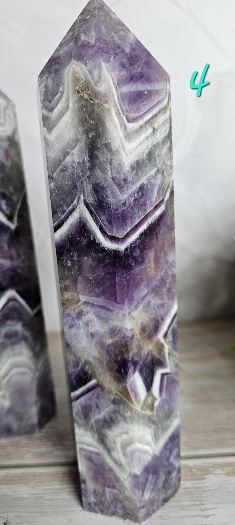 K&ouml;nnte beinhalten: Zwei Amethyst-Obelisken mit poliertem Finish. Der violett-wei&szlig; geb&auml;nderte Stein hat eine kristalline Struktur und ein geometrisches Muster. Die Obelisken sind rechteckig mit einer spitzen Spitze. Die Zahl "4" ist in T&uuml;rkis im Hintergrund geschrieben.