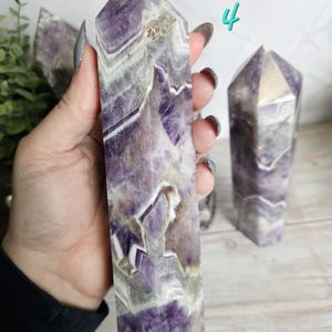 K&ouml;nnte beinhalten: Ein polierter Amethyst-Kristallturm mit spitzer Spitze, der B&auml;nder aus Lila, Wei&szlig; und Grau zeigt. Der Kristall wird in einer Hand gehalten, mit einem weiteren &auml;hnlichen Kristall im Hintergrund. Die Zahl "4" ist in T&uuml;rkis geschrieben.