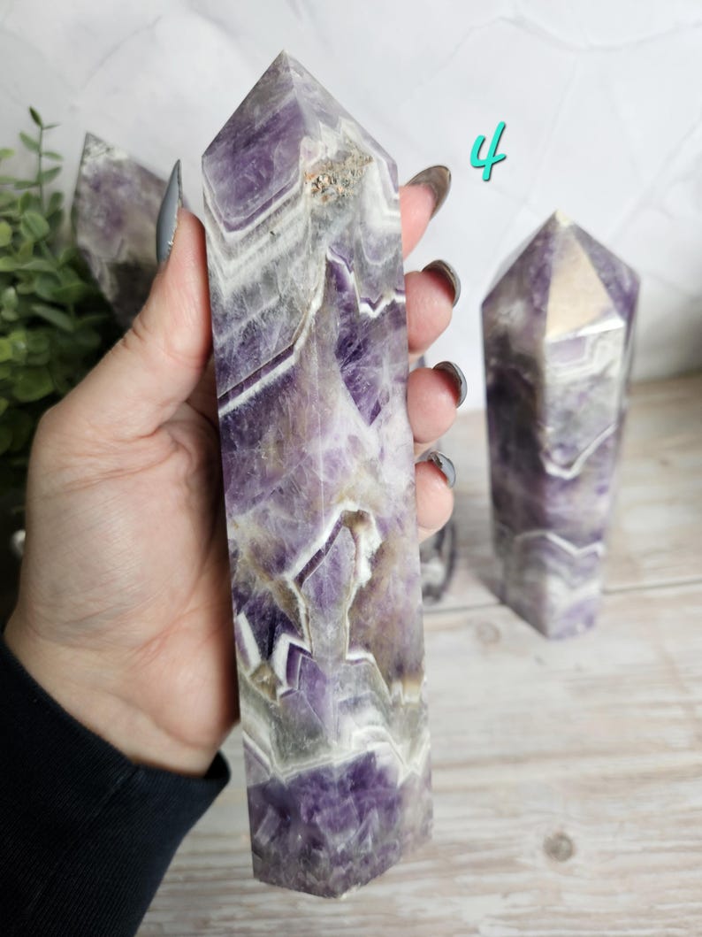 K&ouml;nnte beinhalten: Ein polierter Amethyst-Kristallturm mit spitzer Spitze, der B&auml;nder aus Lila, Wei&szlig; und Grau zeigt. Der Kristall wird in einer Hand gehalten, mit einem weiteren &auml;hnlichen Kristall im Hintergrund. Die Zahl "4" ist in T&uuml;rkis geschrieben.