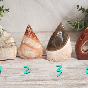 May include: Four polished agate stone sculptures in various colors, including cream, orange, brown, and red. Each stone has a unique shape and pattern, with a geode center visible in one. The stones are numbered 1 through 4.