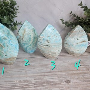 May include: Four polished gemstone sculptures in a teardrop shape. The stones have a mix of blue, white, and tan colors, with a layered appearance. The sculptures are displayed on a light wood surface.