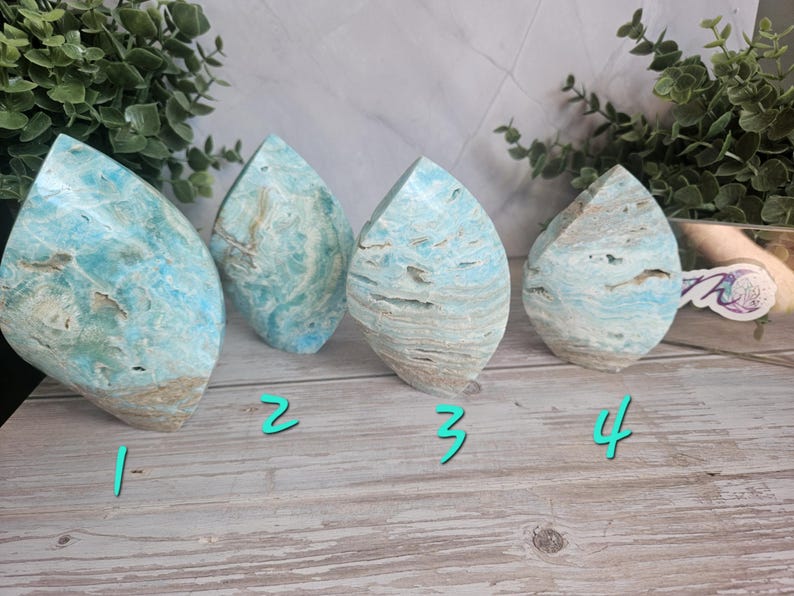 May include: Four polished gemstone sculptures in a teardrop shape. The stones have a mix of blue, white, and tan colors, with a layered appearance. The sculptures are displayed on a light wood surface.