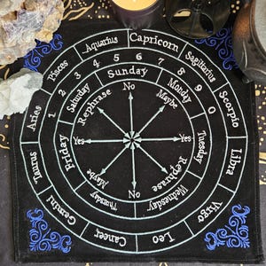 May include: A black square divination cloth featuring white embroidered text including the twelve zodiac signs, days of the week, numbers zero through nine, and the words Yes, No, Maybe, and Rephrase arranged in a circular pattern with arrows.