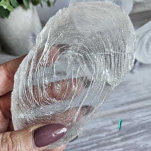 May include: A translucent, oval-shaped selenite crystal slice featuring a distinct, swirling fibrous texture throughout its surface. The mineral specimen is clear with white, vein-like inclusions that create a natural, layered pattern across the stone.