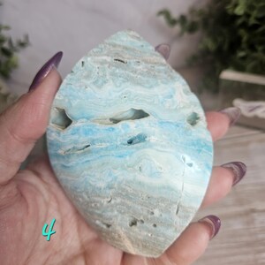May include: A polished, teardrop-shaped gemstone with a blend of light blue, white, and tan hues. The stone has a layered, wave-like pattern and natural cavities. The number "4" is visible in the lower left corner.