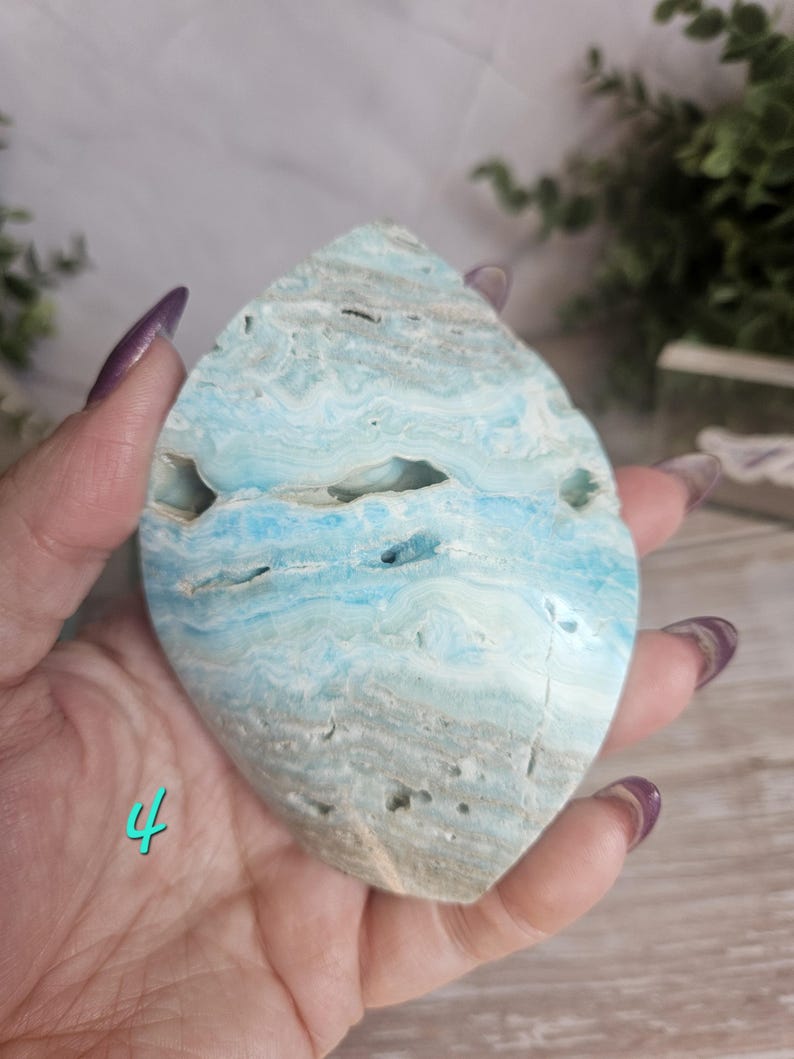 May include: A polished, teardrop-shaped gemstone with a blend of light blue, white, and tan hues. The stone has a layered, wave-like pattern and natural cavities. The number "4" is visible in the lower left corner.