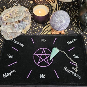 May include: A black rectangular divination mat featuring an embroidered purple pentagram in the center, surrounded by the words Yes, No, Maybe, and Rephrase. A light green crystal pendulum on a silver chain rests on the mat's surface.