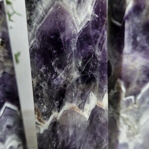 K&ouml;nnte beinhalten: Drei Amethyst-Kristall-Obelisken mit poliertem, facettiertem Design. Die Kristalle zeigen eine Mischung aus tiefen Lila-, Wei&szlig;- und Graut&ouml;nen. Die Obelisken stehen aufrecht und pr&auml;sentieren ihre geometrische Form und nat&uuml;rlichen Muster.