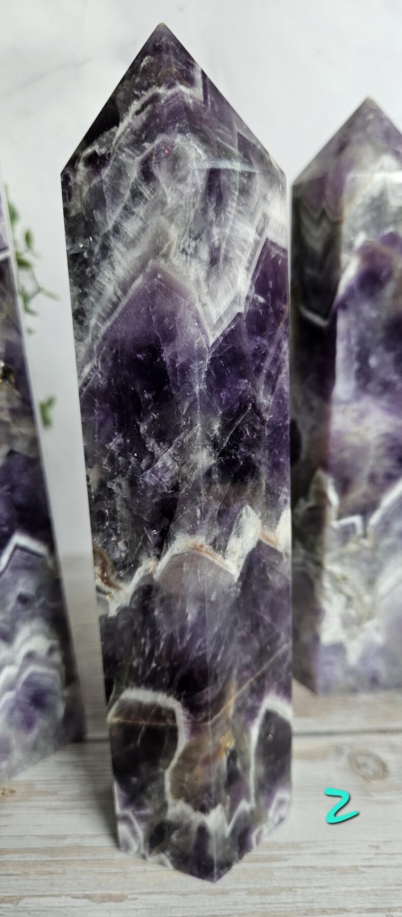K&ouml;nnte beinhalten: Drei Amethyst-Kristall-Obelisken mit poliertem, facettiertem Design. Die Kristalle zeigen eine Mischung aus tiefen Lila-, Wei&szlig;- und Graut&ouml;nen. Die Obelisken stehen aufrecht und pr&auml;sentieren ihre geometrische Form und nat&uuml;rlichen Muster.