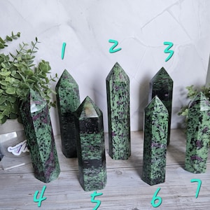 May include: Seven green and red zoisite crystal towers with pointed tops. The crystals have a speckled appearance, with a mix of green and red hues. The crystals are arranged on a light-colored surface, with greenery in the background.