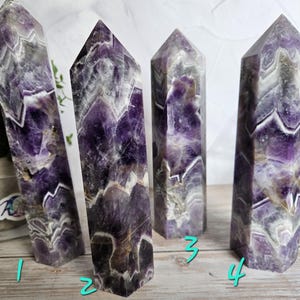 K&ouml;nnte beinhalten: Vier Amethyst-Kristall-Obelisken mit polierter Oberfl&auml;che. Die Kristalle zeigen eine Mischung aus tiefen Lila-, Wei&szlig;- und Graut&ouml;nen mit sichtbaren Chevron-Mustern. Die Obelisken stehen aufrecht und pr&auml;sentieren ihre geometrischen Formen und ihre nat&uuml;rliche Sch&ouml;nheit.
