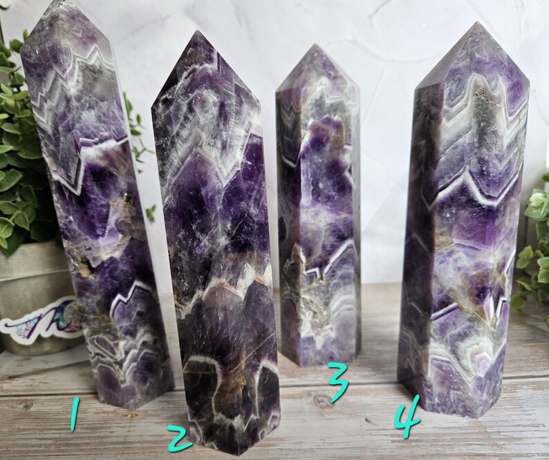 K&ouml;nnte beinhalten: Vier Amethyst-Kristall-Obelisken mit polierter Oberfl&auml;che. Die Kristalle zeigen eine Mischung aus tiefen Lila-, Wei&szlig;- und Graut&ouml;nen mit sichtbaren Chevron-Mustern. Die Obelisken stehen aufrecht und pr&auml;sentieren ihre geometrischen Formen und ihre nat&uuml;rliche Sch&ouml;nheit.