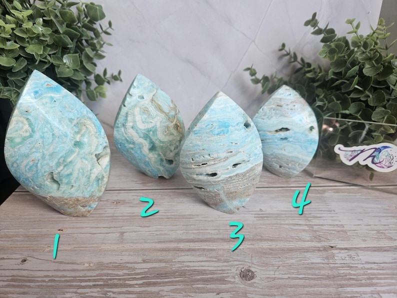 May include: Four teardrop-shaped, polished stone sculptures. The stones have a light blue and beige color scheme with a marbled appearance. The sculptures are displayed on a light wood-grain surface with greenery in the background. Each stone has a unique pattern.