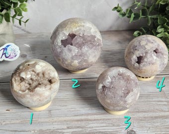 May include: Four amethyst crystal spheres on cream-colored stands. The spheres vary in size and have a mix of light purple and white coloring. Some spheres have open geodes, revealing the crystal interiors. The spheres are displayed on a light wood surface.