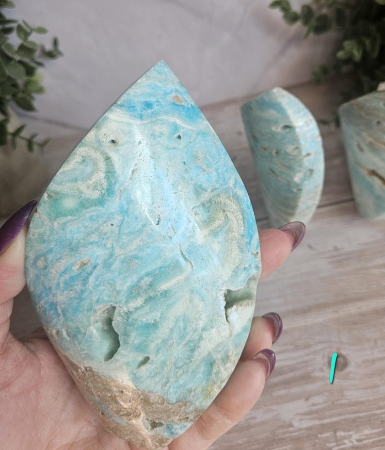 May include: A large, teardrop-shaped blue aragonite crystal with white and tan veining. The polished stone has natural holes and a rough, unpolished base. Two additional aragonite pieces are visible in the background.