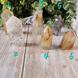 May include: Six clear quartz crystal points with brown and gold inclusions. The points are arranged in a row on a wooden surface.