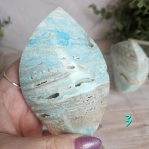 May include: A large, teardrop-shaped piece of polished aragonite in shades of blue, white, and tan. The stone has a layered appearance with natural holes and textures. A smaller, similar piece is visible in the background.