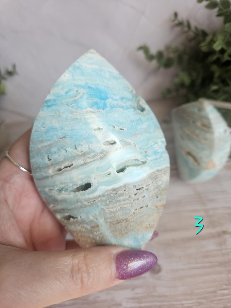 May include: A large, teardrop-shaped piece of polished aragonite in shades of blue, white, and tan. The stone has a layered appearance with natural holes and textures. A smaller, similar piece is visible in the background.