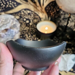 May include: A small, matte black bowl with a curved, undulating rim and a circular base. The bowl has a smooth, solid-colored exterior and a deep, open interior, held in a hand to display its size and shape against a dark, patterned backdrop.