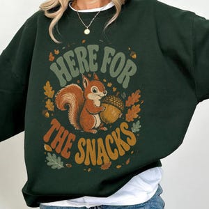 Puede incluir: Una sudadera de cuello redondo en color verde bosque oscuro con un estampado gráfico de una ardilla marrón sosteniendo una bellota grande. El texto "HERE FOR THE SNACKS" está impreso en una fuente retro curva, rodeado de hojas de roble naranjas y verdes.